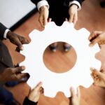 Team members place hands around piece of paper in the shape of a cog, symbolizing successful change management and project success.